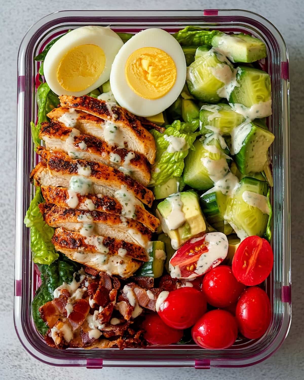 Cobb salad meal prep box filled with grilled chicken, avocado, tomatoes, cucumber, bacon, and a hard-boiled egg.