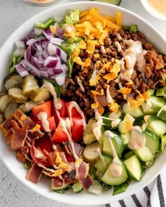 Cheeseburger bowl meal prep with seasoned ground beef, lettuce, tomatoes, pickles, red onion, bacon, cheese.